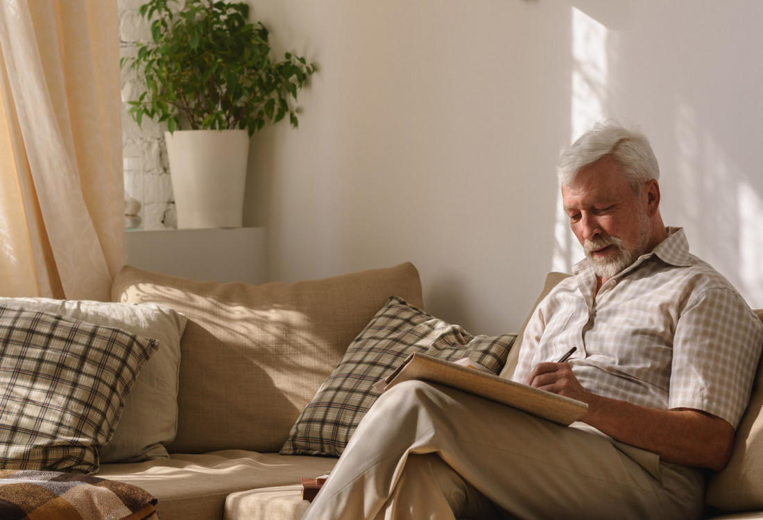 Ein alter Mann sitzt auf einem Sofa und schreibt in ein Notizbuch.